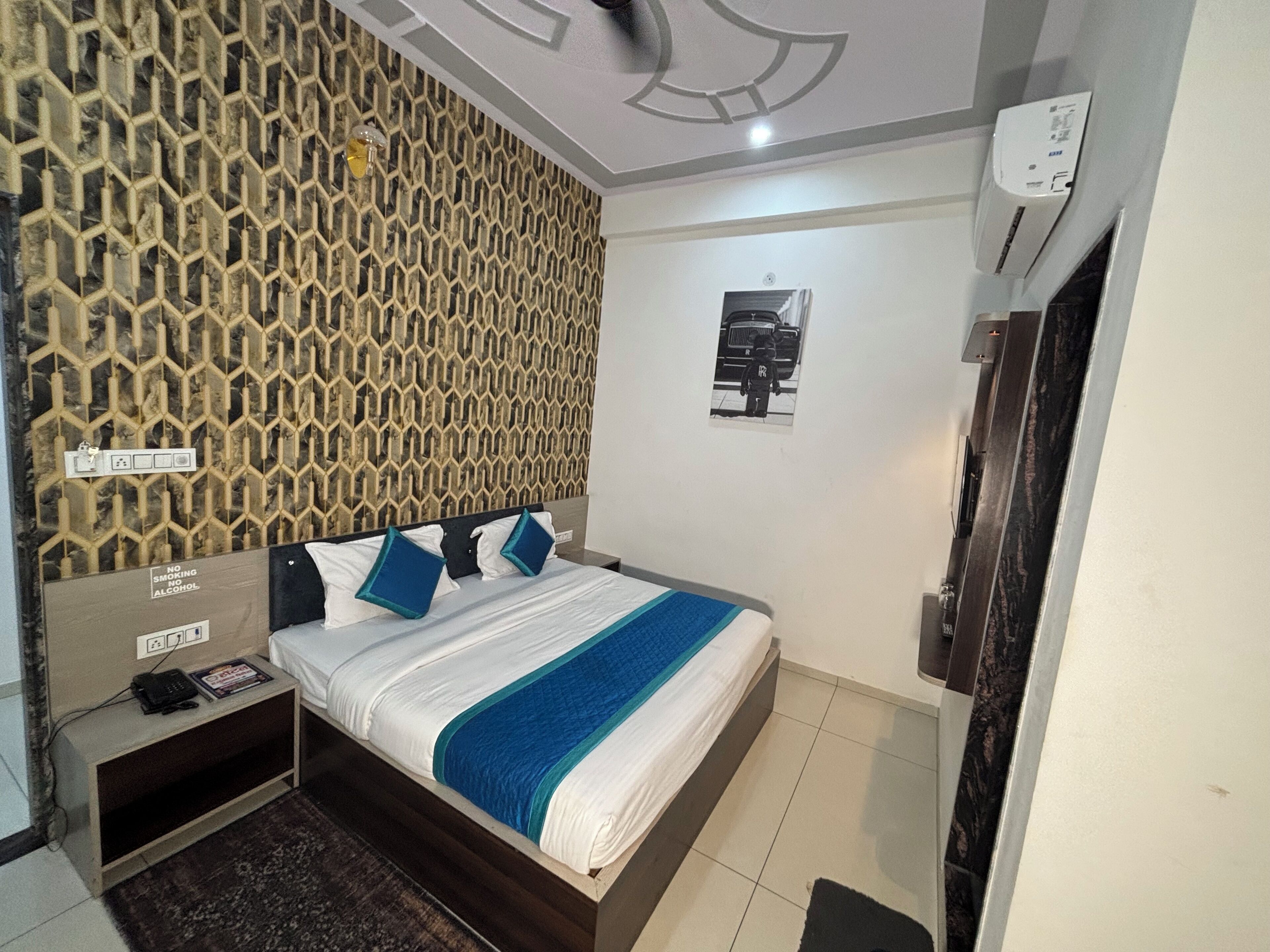 A hotel room with a patterned wall, a bed with blue and white bedding, a bedside table with a phone, and a wall-mounted air conditioning unit.