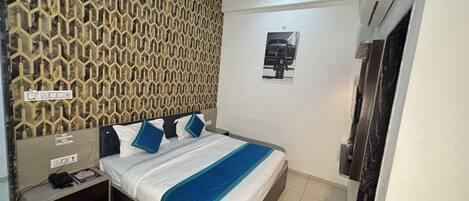 A hotel room with a patterned wall, a bed with blue and white bedding, a bedside table with a phone, and a wall-mounted air conditioning unit.