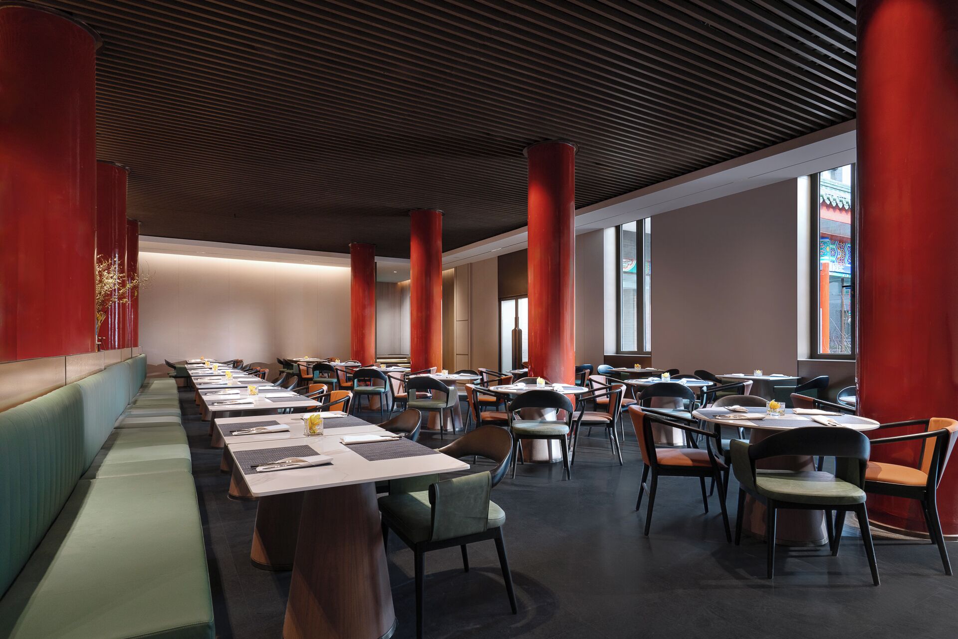 Een moderne eetruimte met lange tafels en stoelen, grote ramen en rode zuilen.