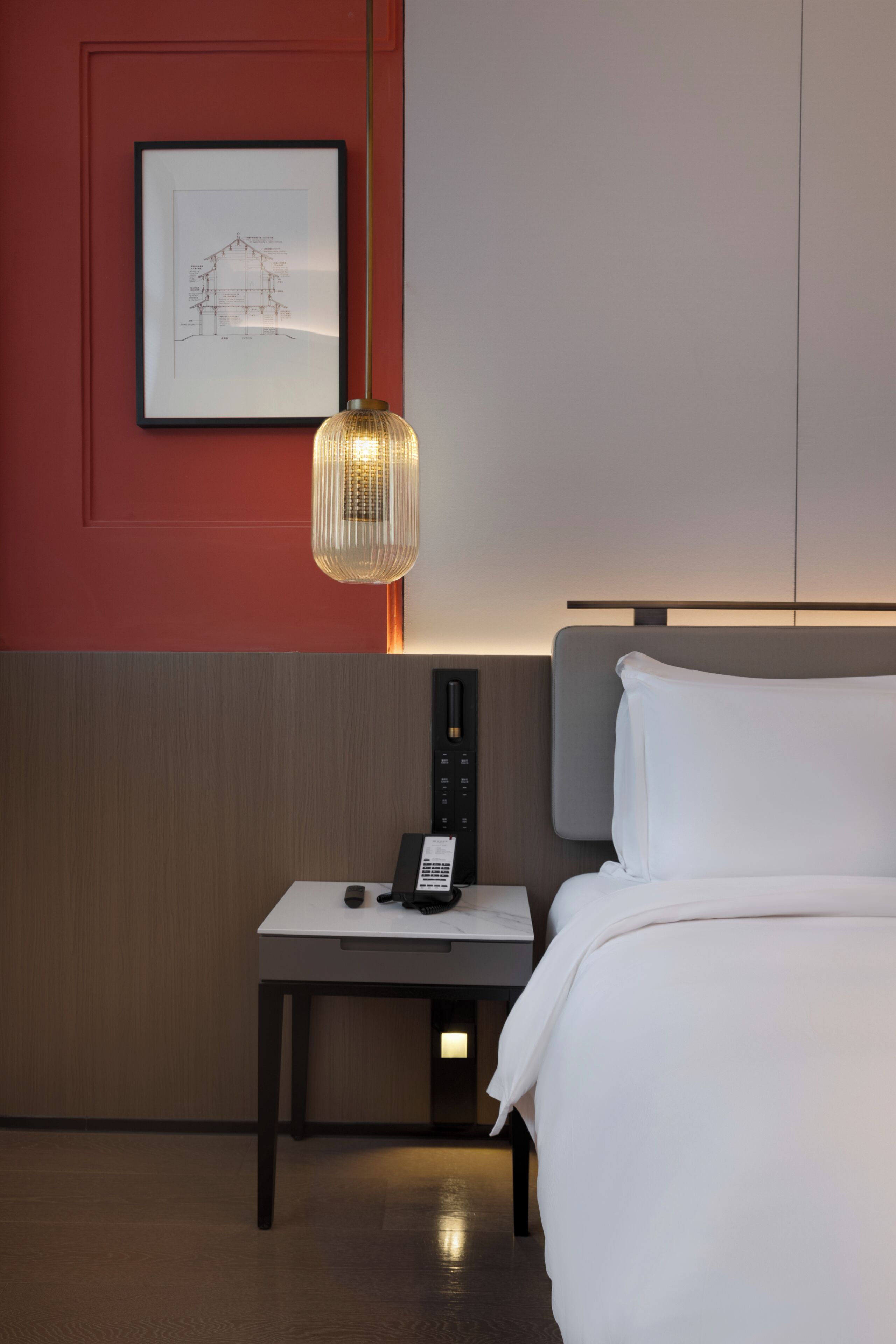 A hotel room with a bed, bedside table, lamp, and a framed architectural drawing on the wall.