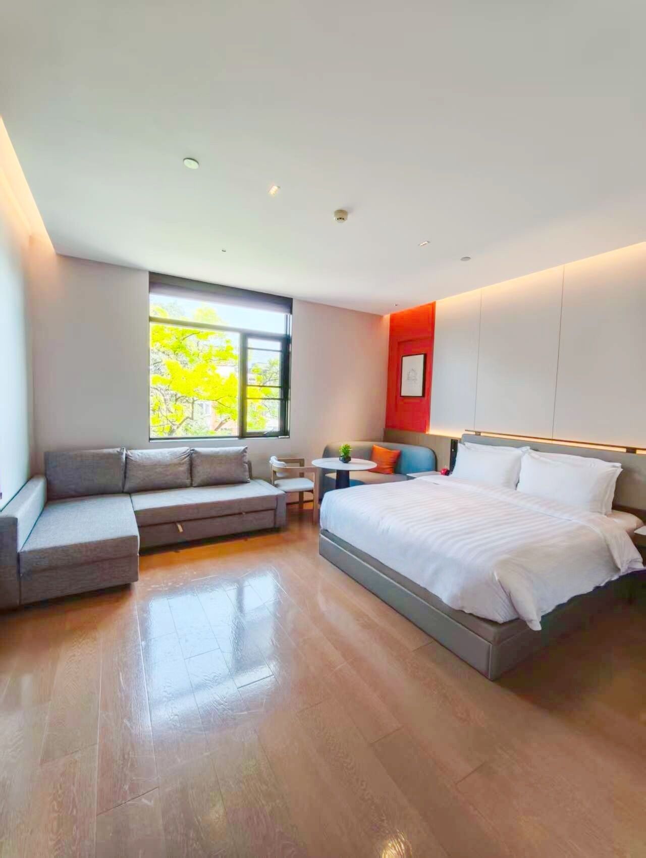 A modern bedroom with a bed, a sofa, a small table, and a window with a view of trees.