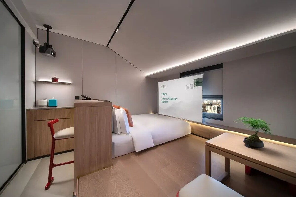 Une chambre moderne et compacte, comprenant un lit, un bureau, une chaise, une petite table avec une plante et une fenêtre donnant sur la ville.