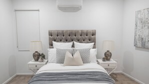 Ein Schlafzimmer mit einem gebügelten Kopfteil, zwei Nachttischen mit Lampen, einem großen Bett mit einer grauen und weißen Musterdecke sowie einer Wand-Klimaanlage.