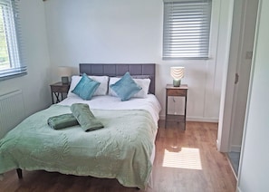 A bedroom with a bed, pillows, a bedside table with a lamp, and a window with blinds. - The Country oak is a 2 double bedroom lodge with en suites to both rooms. (Langworth)