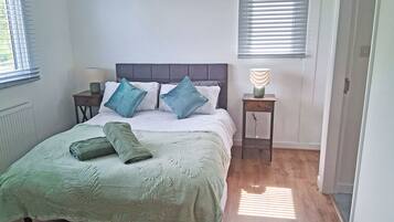 A bedroom with a bed, pillows, a bedside table with a lamp, and a window with blinds.