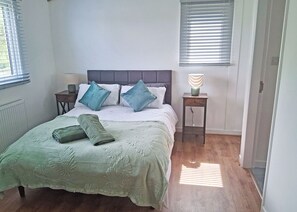 A bedroom with a bed, pillows, a bedside table with a lamp, and a window with blinds.