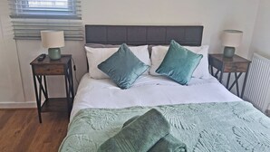 Una cama con cabecera gris, dos mesitas de noche con lámparas, y un ventanal con persianas.