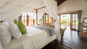 A spacious bedroom with a large bed, white bedding, and a canopy. There are wooden floors and a view of the outdoors through large windows. - Uma Nawasena Ubud (Ubud)
