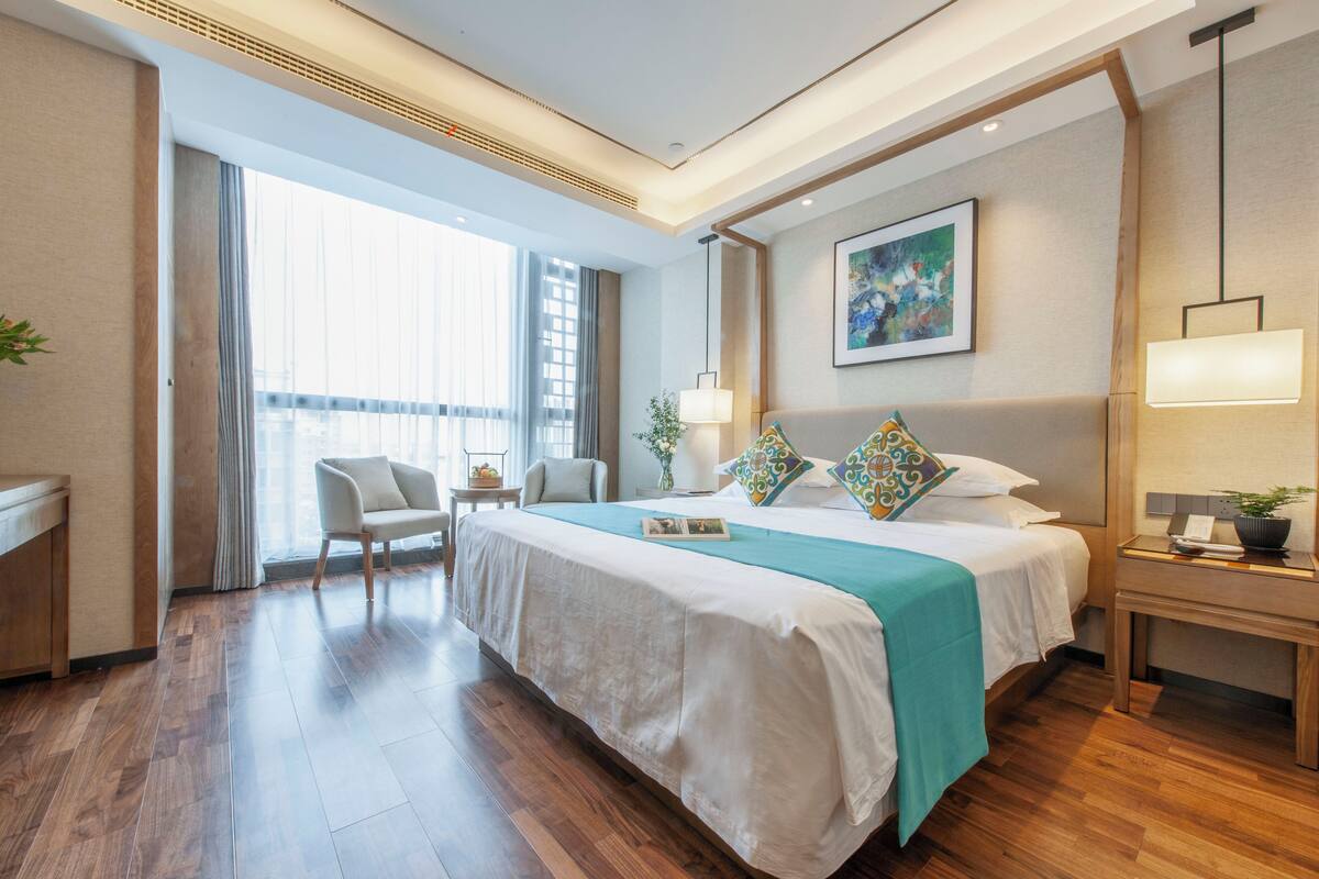 A modern hotel room with a large bed, a desk, a chair, a bedside table with a lamp, and a window with curtains.