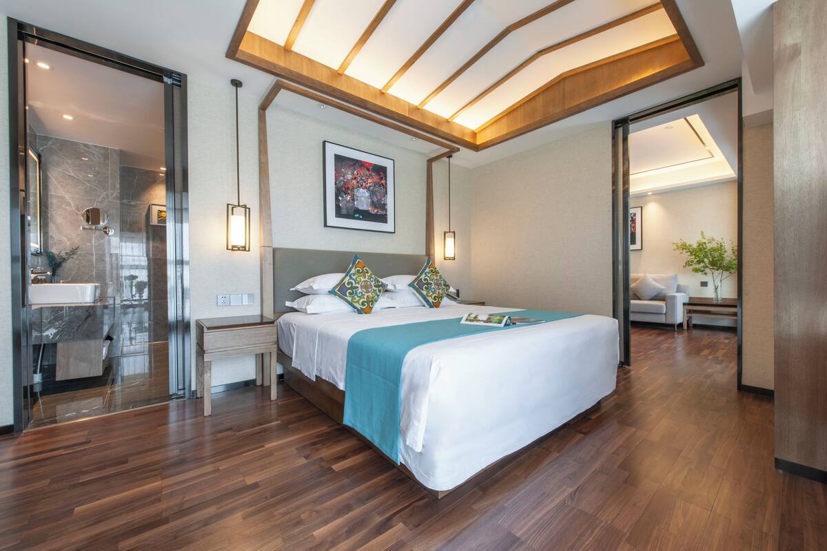 A modern hotel room with a large bed, wooden flooring, a bathroom with a glass shower, and a dining area with a table and chairs.