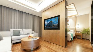 Ett modernt hotellrum med trägolv, ett runt soffbord, en soffa, en säng, en tv som är monterad på en träpanel och ett sängbord med en lampa.