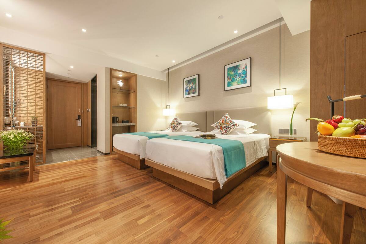 A hotel room with two beds, wooden flooring, a dining area with a table and chairs, a fruit basket, and artwork on the walls.