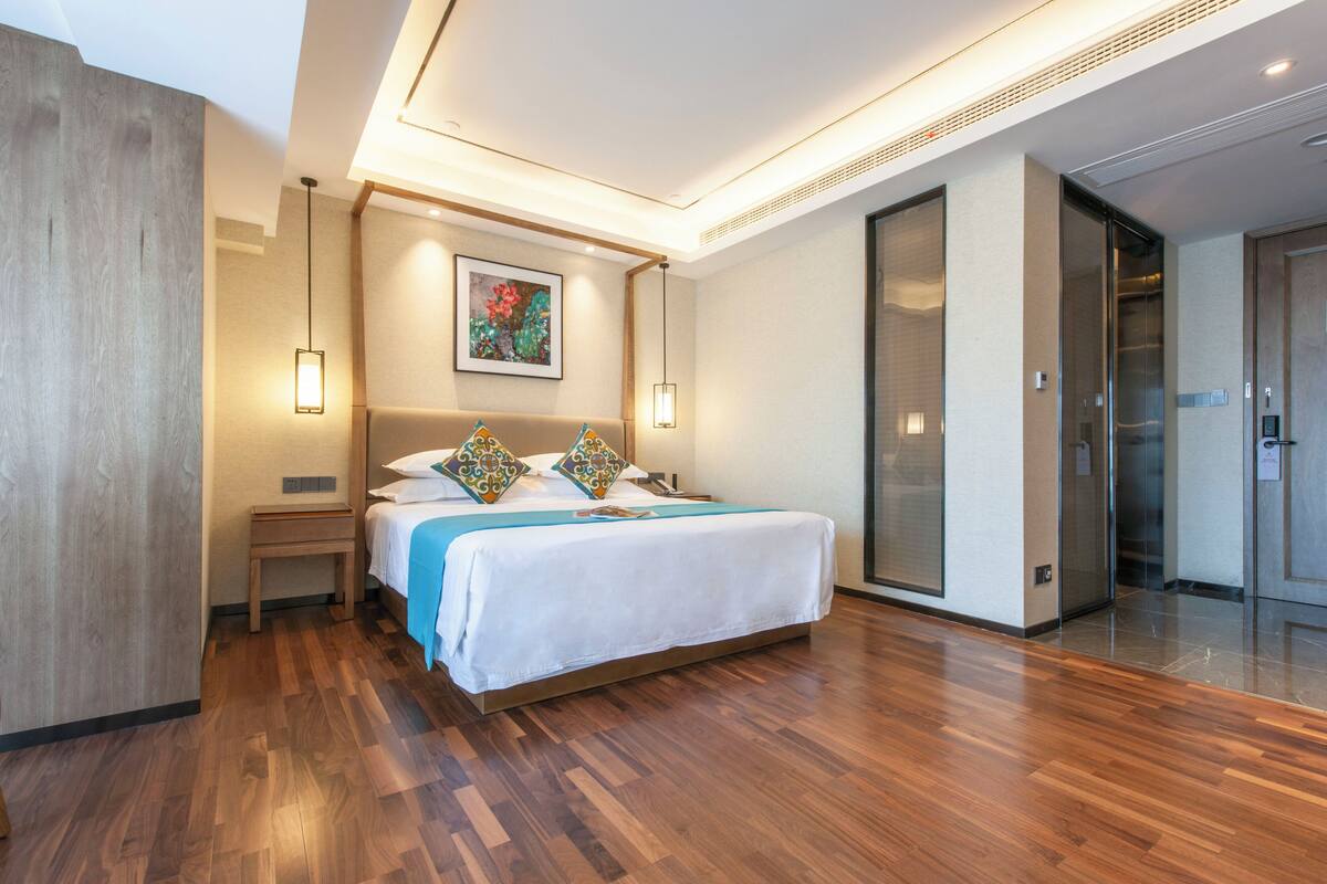 A modern hotel room with a wooden floor, a large bed with patterned pillows, a painting on the wall, and a sliding door closet.