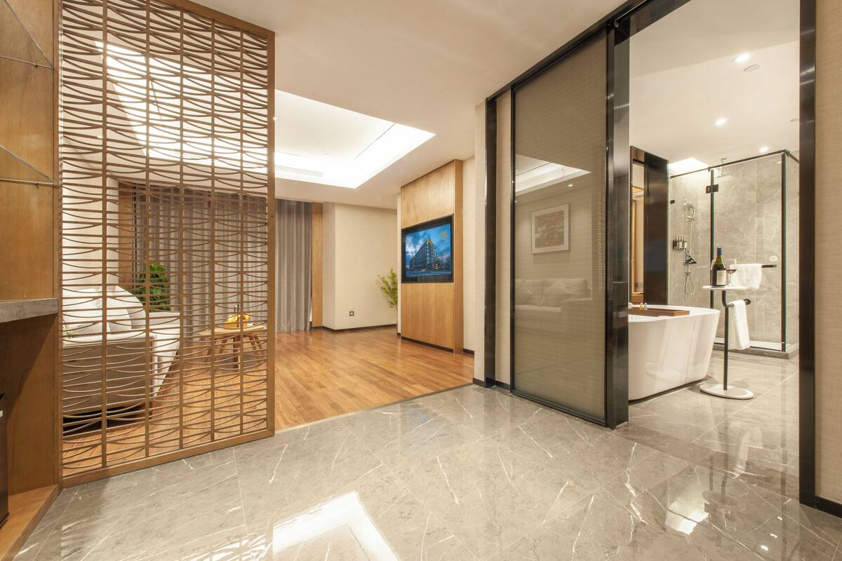A modern hotel room with a wooden partition, a large window, a bathroom with a glass shower, and a flat-screen TV.