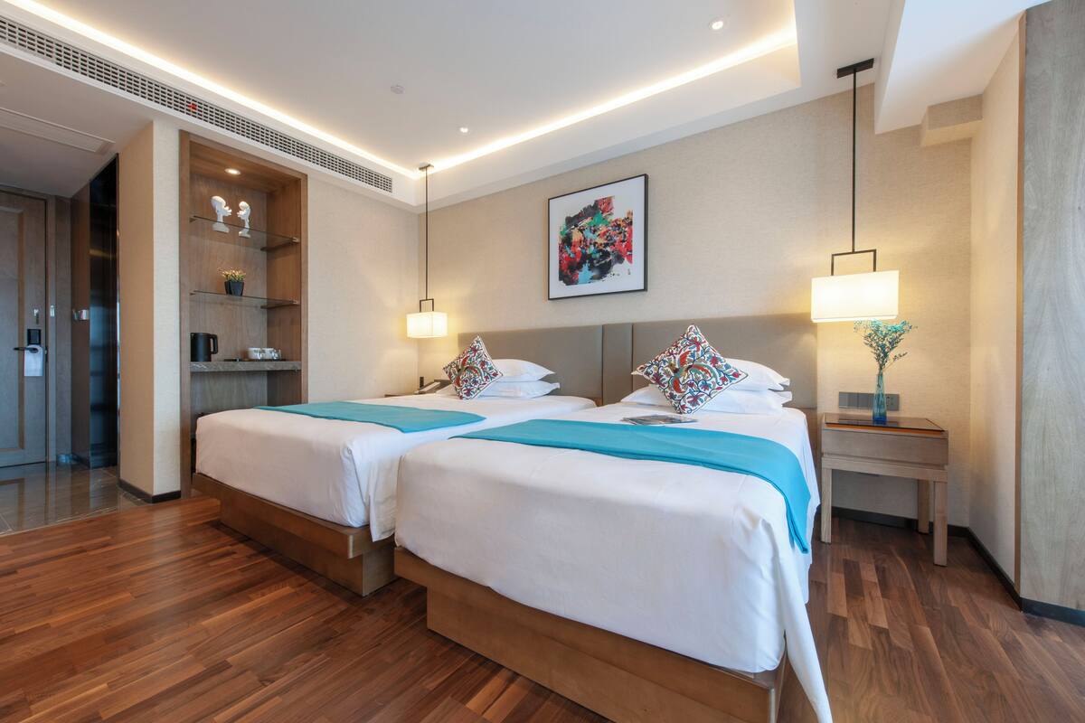 A hotel room with two beds, a small table, a nightstand, a wall-mounted artwork, and a built-in shelf with decorative items.