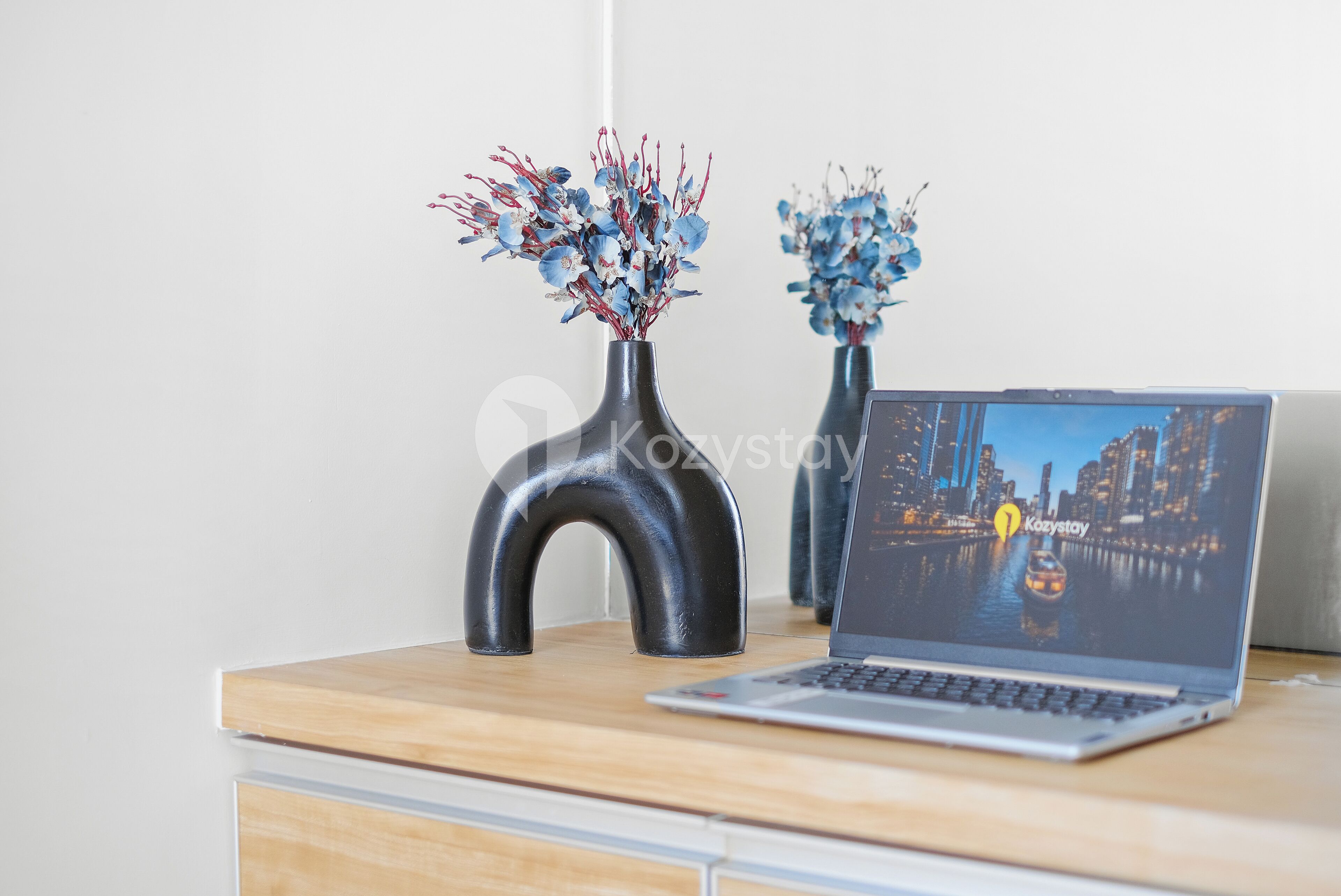 A laptop on a wooden desk with two vases of flowers and a black, curved object.