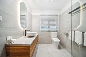 Een moderne badkamer met een grote, ovale spiegel, een houten wastafelmeubel, een witte wasbak, een toilet en een douchecabine met glazen wanden.