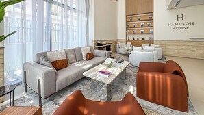 A modern hotel lobby with a grey sofa, a coffee table, and a seating area with orange chairs. A bookshelf with framed pictures is on the wall, and the hotel name 'Hamilton House' is visible.