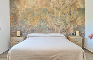 Un dormitorio con cabecera de piedra, una cama con cubrecama blanco, dos mesitas de noche con lámparas y mesitas auxiliares de madera.