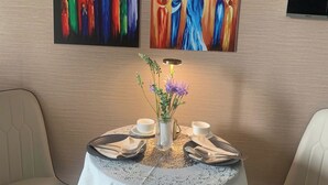 Un comedor con una mesa redonda preparada para dos personas, un florero con flores y cuadros en la pared.