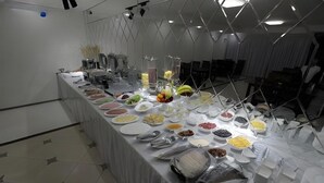 A buffet setup with various food items and utensils on a long table in a modern room with geometric ceiling design. - Sharq Hotel (Urgench)