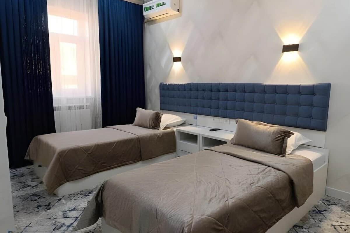 A hotel room with two beds, a large window with curtains, a wall-mounted air conditioner, and a white desk.