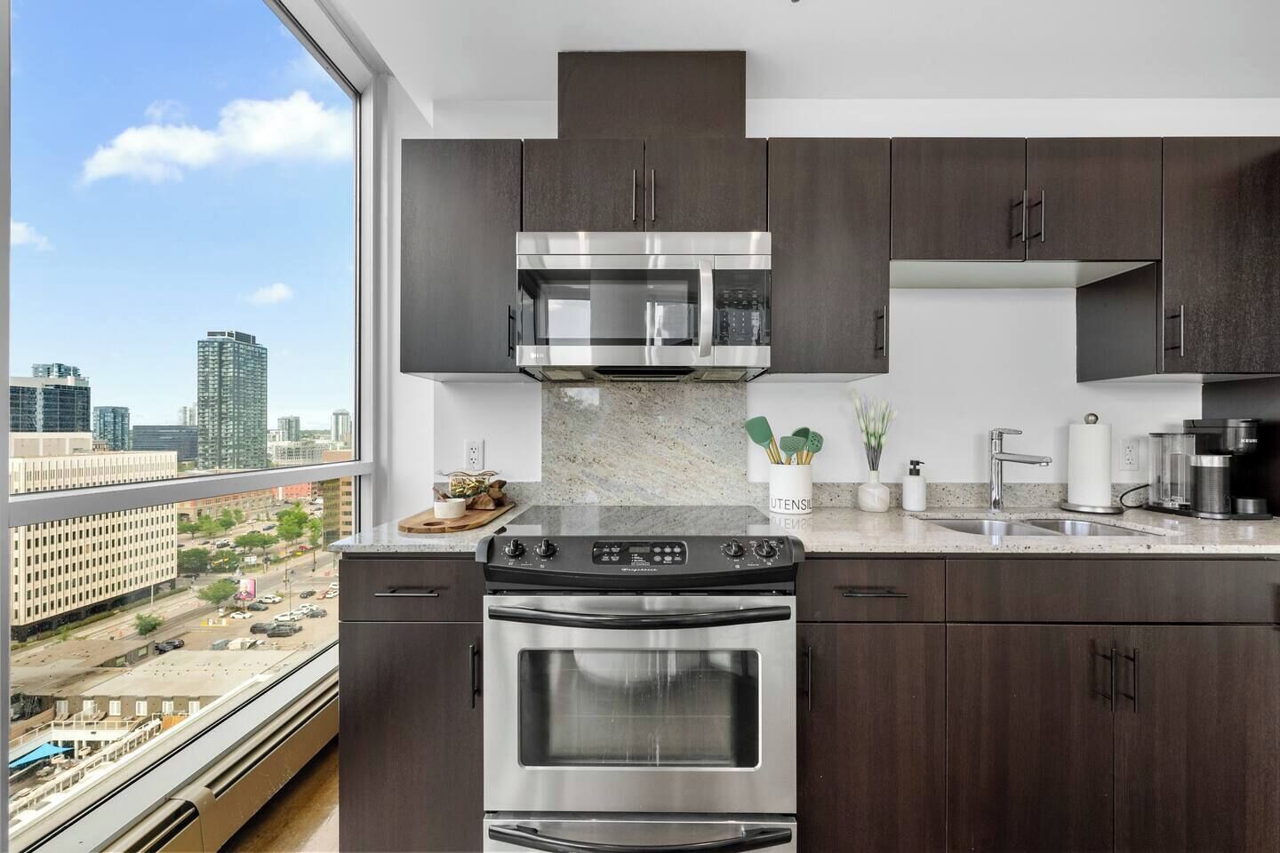 A modern kitchen with dark wood cabinets, stainless steel appliances, and a large window offering a city view.