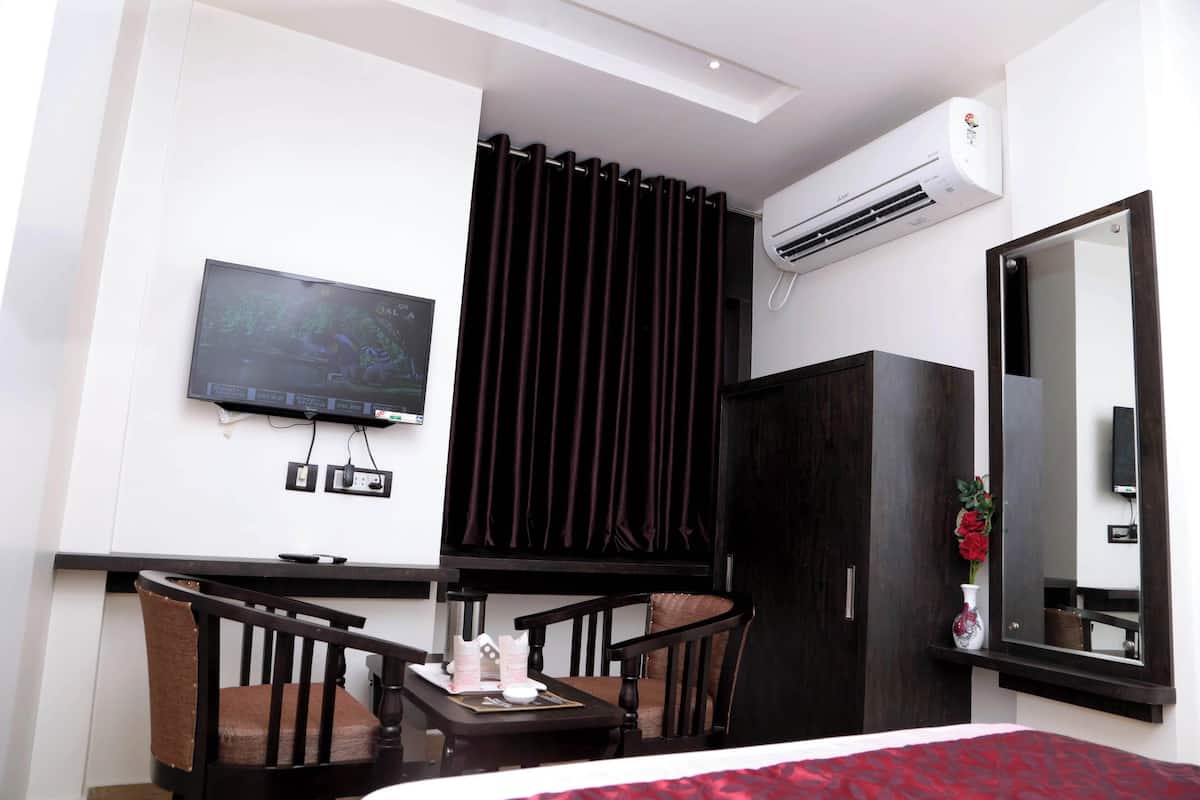 A hotel room with a bed, a TV mounted on the wall, a dark wooden wardrobe, a small table with a chair, and an air conditioning unit.