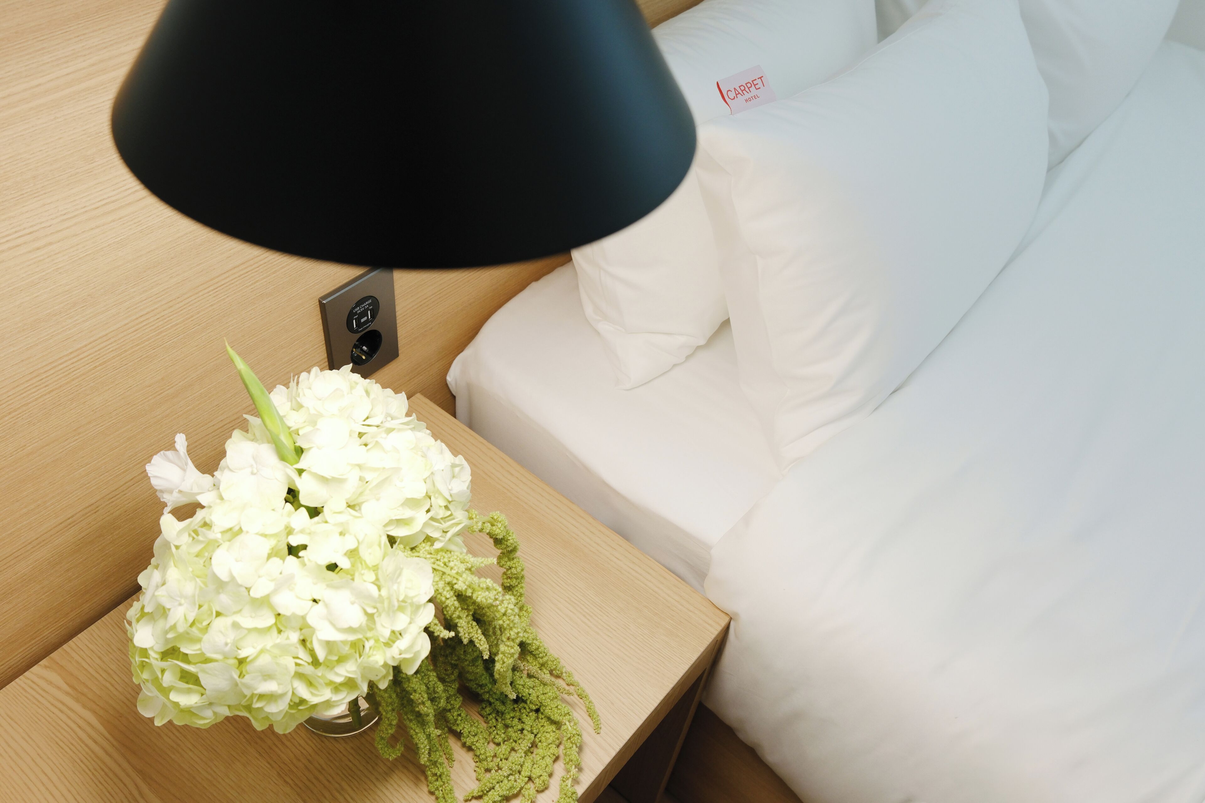 Una cama bien hecha con ropa blanca y una mesita de noche con una lámpara negra y un florero con flores blancas.