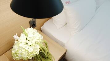 Una cama bien hecha con ropa blanca y una mesita de noche con una lámpara negra y un florero con flores blancas.