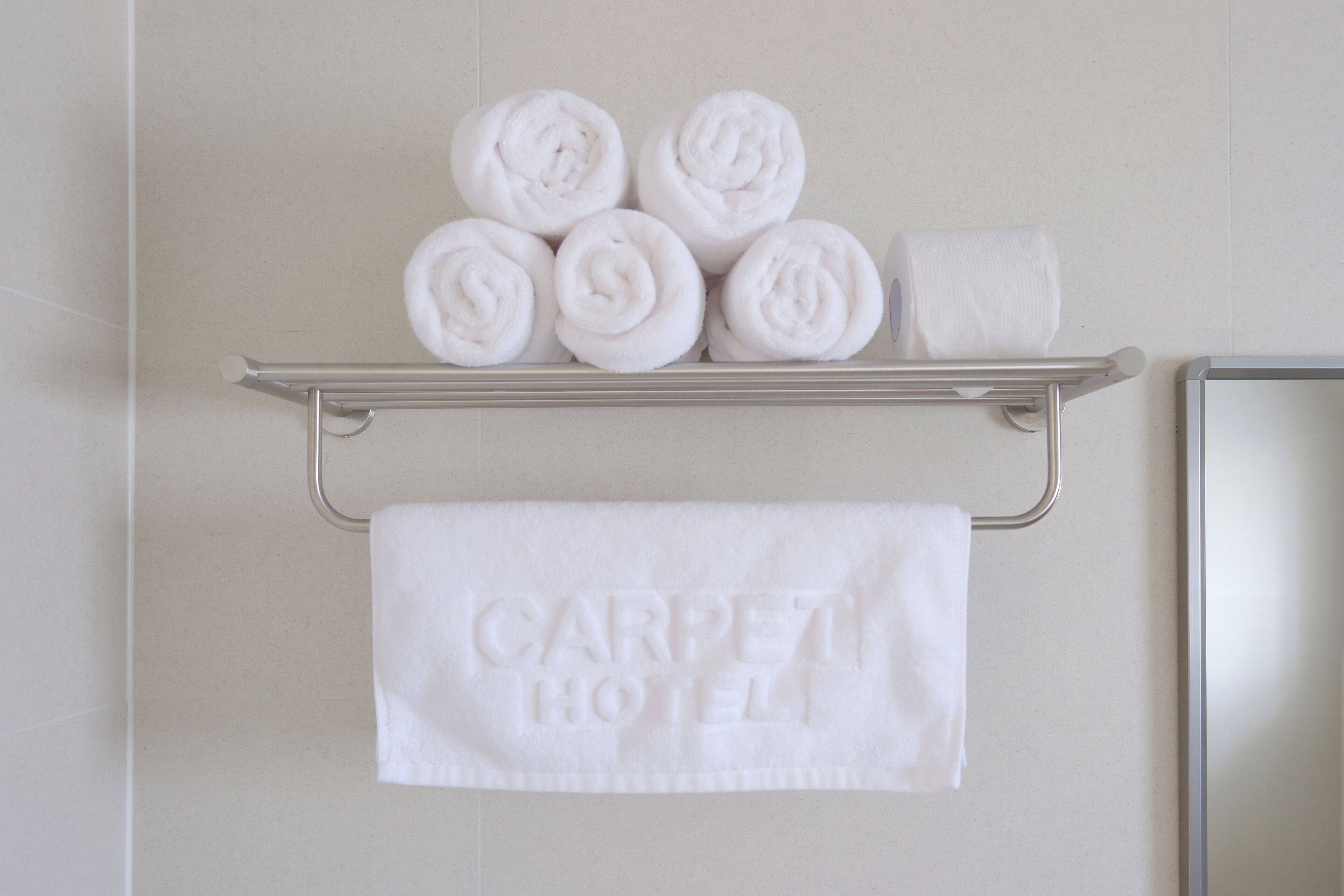 Un baño con un toallero que sostiene toallas enrolladas y una toalla con el grabado 'CARPET HOTEL'.