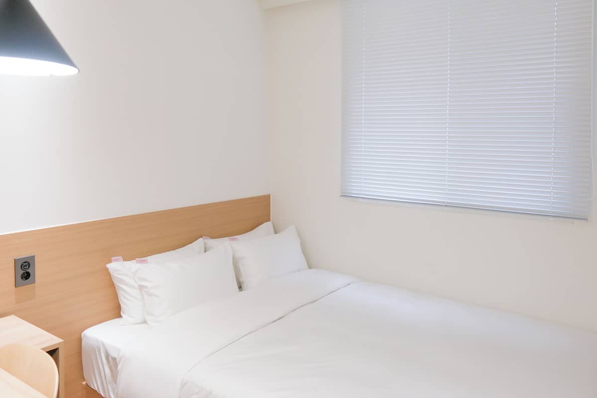 A hotel room with a bed, a nightstand, a lamp, and a window with blinds.