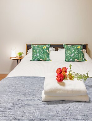 A neatly made bed with white linens, two green floral pillows, a white towel, and a small bedside table with a lamp and a plant. - ideas ASAKUSA (Tokyo)