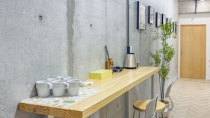 Un'area bar stretta e allungata con un bancone in legno, diverse tazze bianche, una scatola gialla e una macchina per il caffè. Sono presenti sedie in metallo e una pianta in vaso.