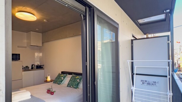 Una camera d'albergo moderna con una porta scorrevole su un balcone, un letto con coperta blu, un angolo cottura e una lavagna con del testo.