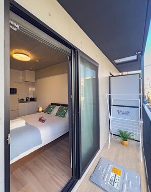 A modern hotel room with a sliding door to a balcony, a bed with a blue blanket, a small kitchen area, and a whiteboard with text. - ideas ASAKUSA (Tokyo)