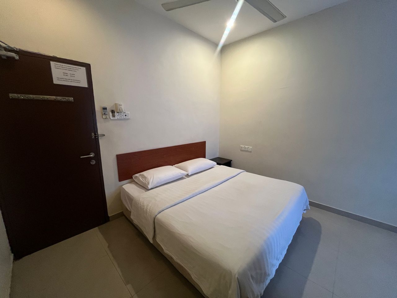Habitación de hotel con cama, puerta de madera, mesita de noche y lámpara fijada a la pared.