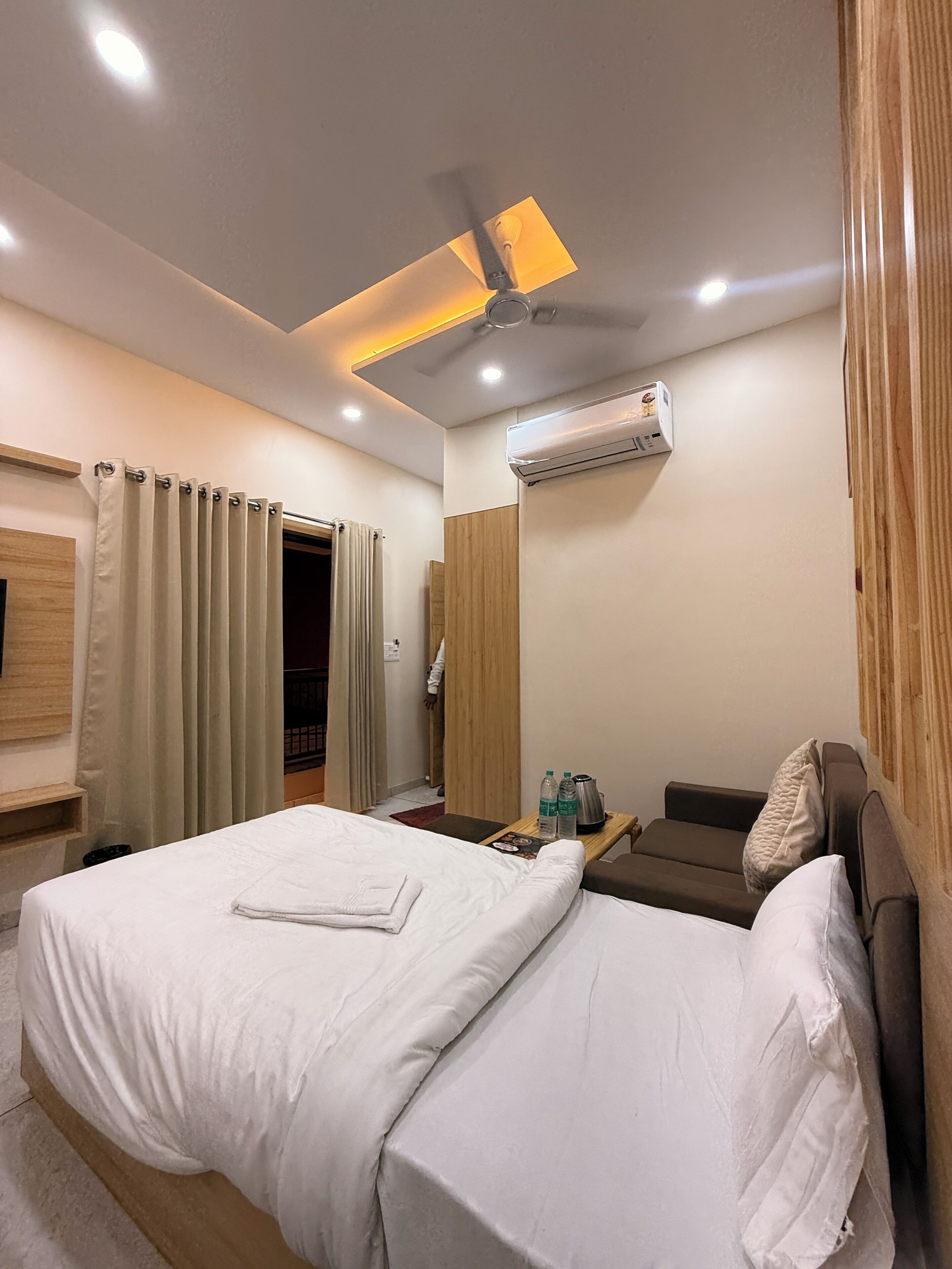 Habitación de hotel con cama, mesita de noche con hervidor y botellas de agua, unidad de aire acondicionado, ventilador de techo y cabecero de madera.