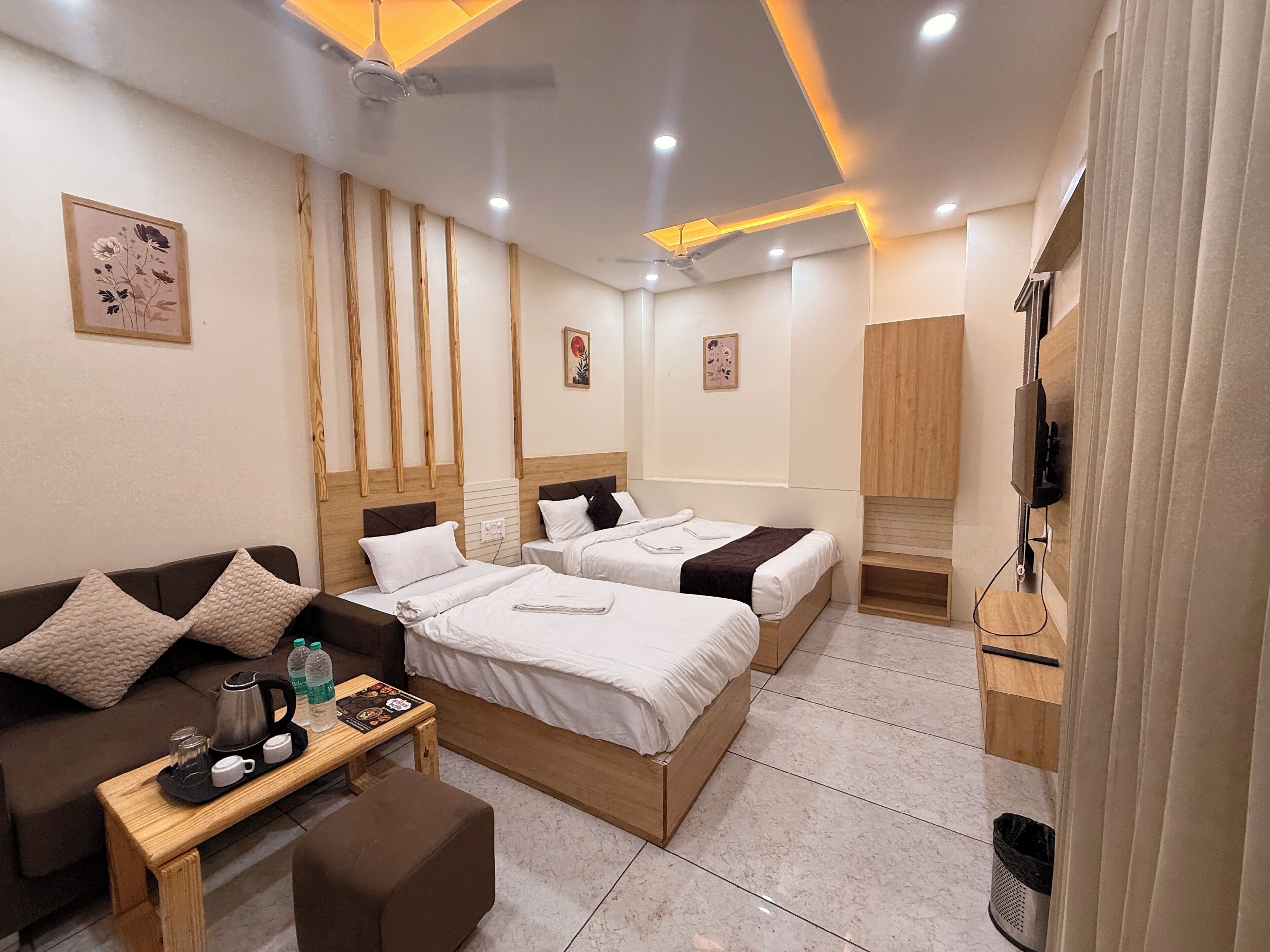 A hotel room with two beds, a sofa, a small table with a kettle and cups, and a TV mounted on the wall.