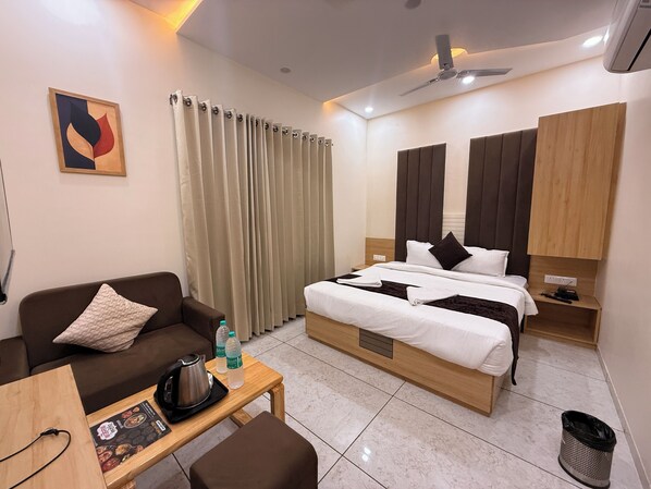 Una habitación de hotel con cama, sofá, una mesita con tetera y botellas, un cesto de basura y un ventilador de techo.