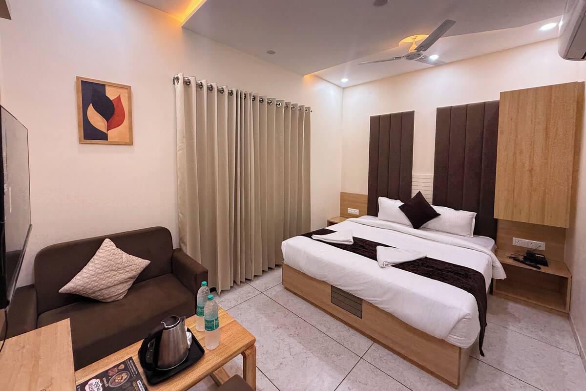 Quarto de hotel com cama, sofá, mesa de centro com chaleira e garrafas, e um quadro na parede.