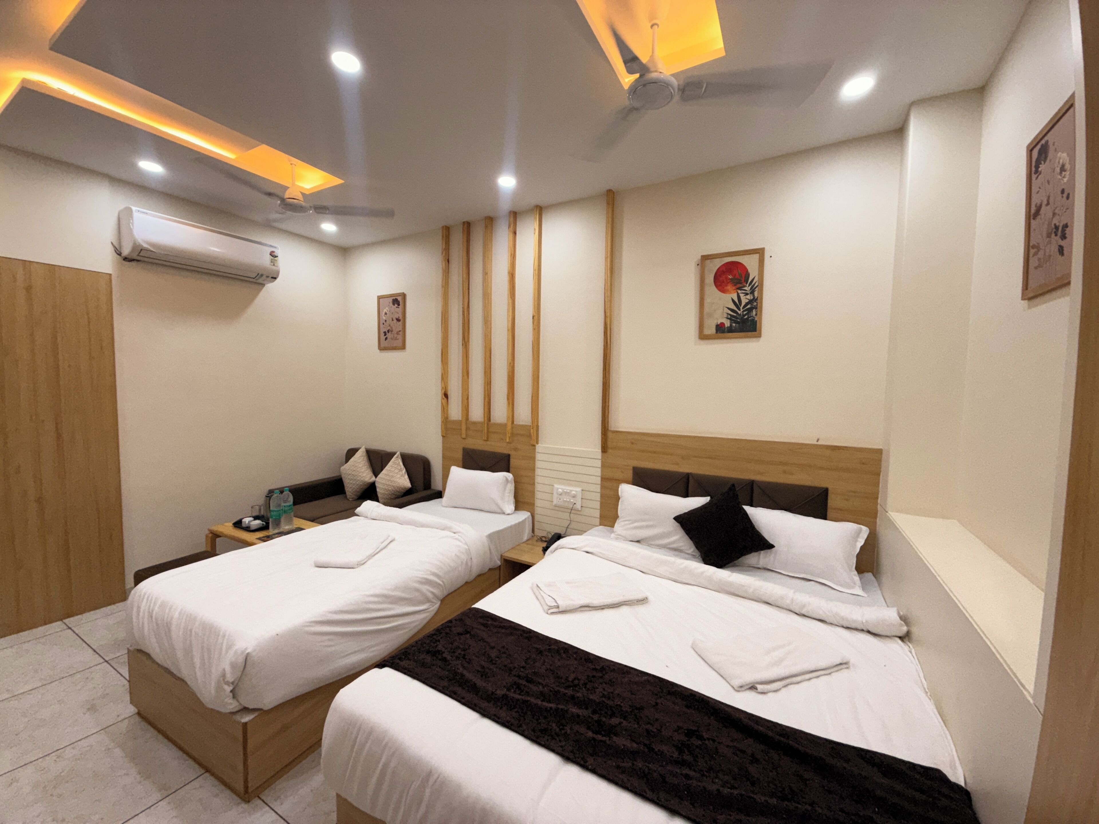 Habitación de hotel con dos camas, cabecera de madera, una mesita con una silla, unidad de aire acondicionado y una obra de arte en la pared.