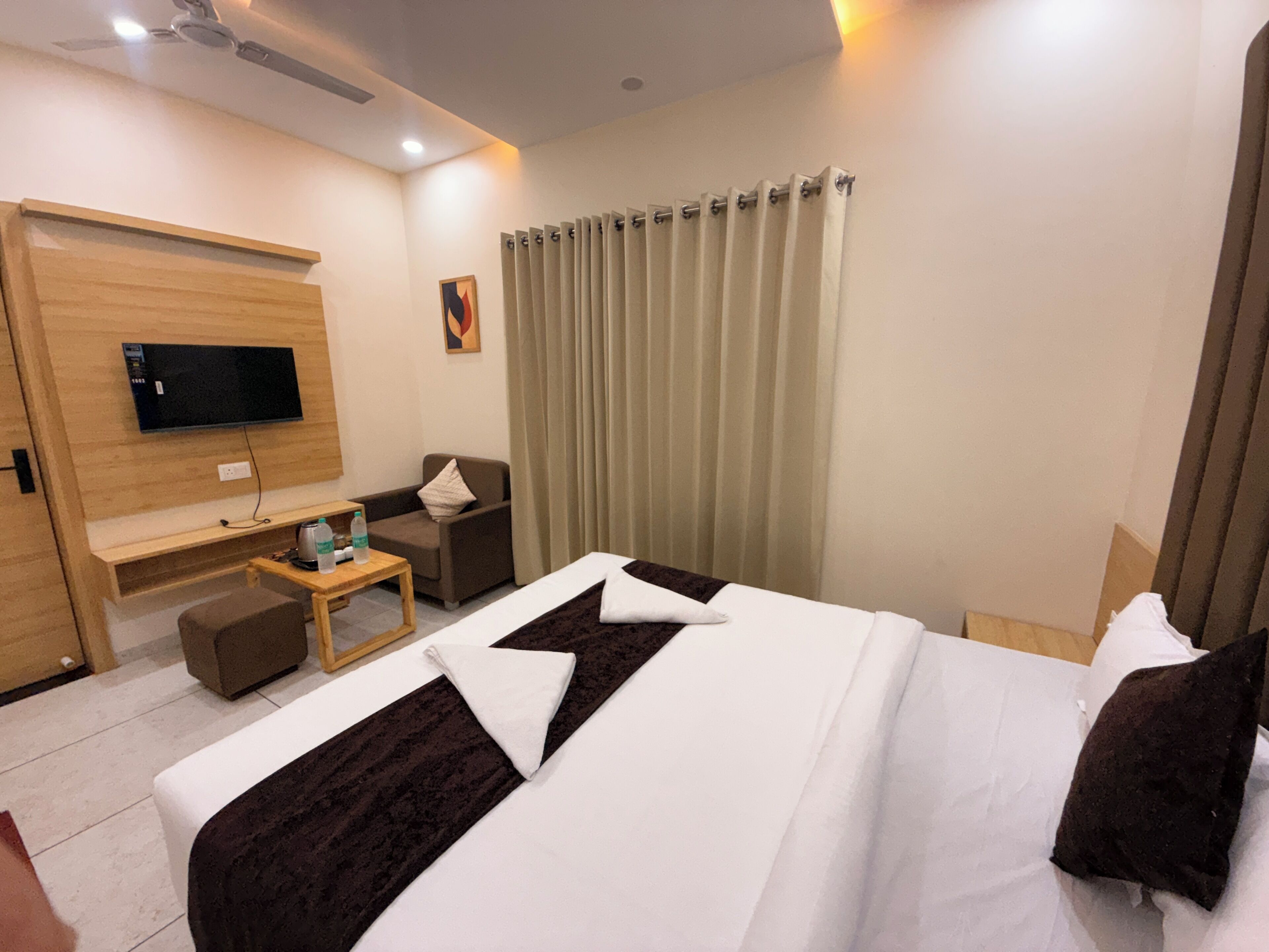 A hotel room with a bed, a TV mounted on the wall, a desk with a chair, and a window with curtains.