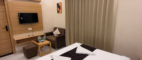 A hotel room with a bed, a TV mounted on a wooden panel, a small table with a bottle, a chair, and a curtain.