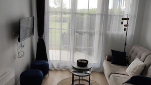 A living room with a sofa, a small table with a bowl, a TV mounted on the wall, and a large window with sheer curtains. - Apartment -2 bedrooms, ground floor Port Bourgenay village with swimming pool access (Talmont-Saint-Hilaire)