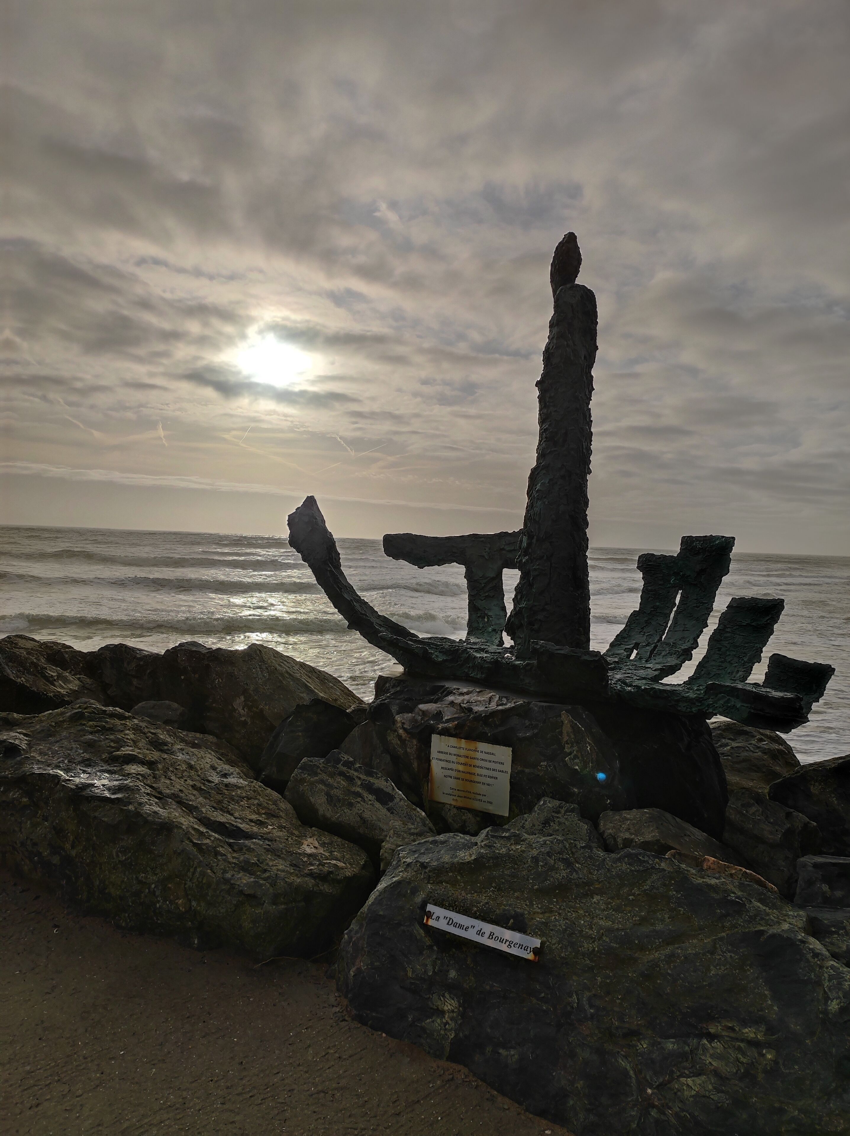 A rocky coastal landscape with a large, weathered structure, possibly a historical monument, and a sign indicating "La Dame de Bourgenay."