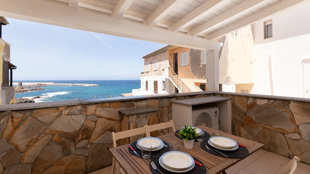 A terrace with stone walls, a wooden table set for dining, and a view of the sea and buildings.