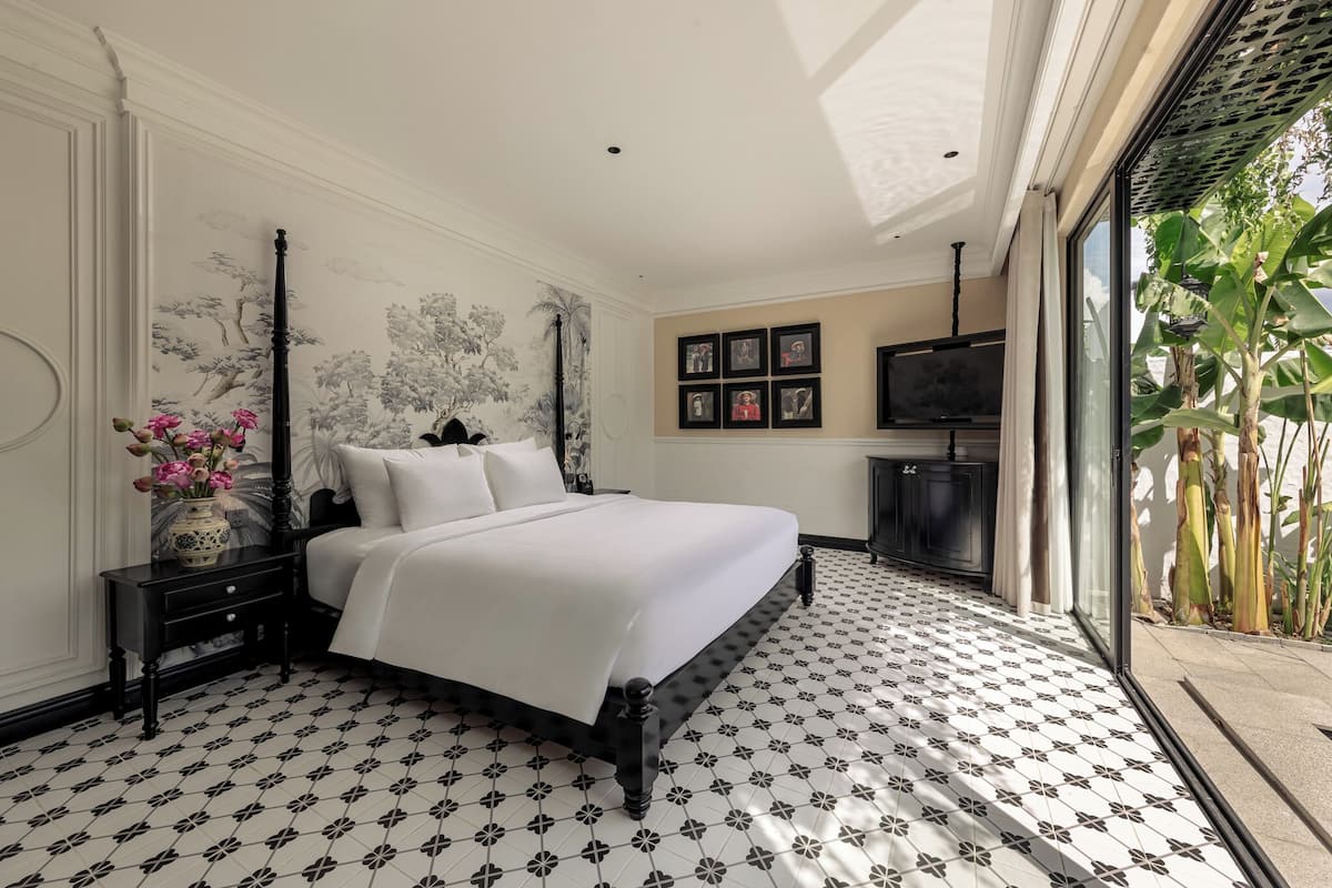 A bedroom with a bed, nightstand, and a large window with a view of plants.