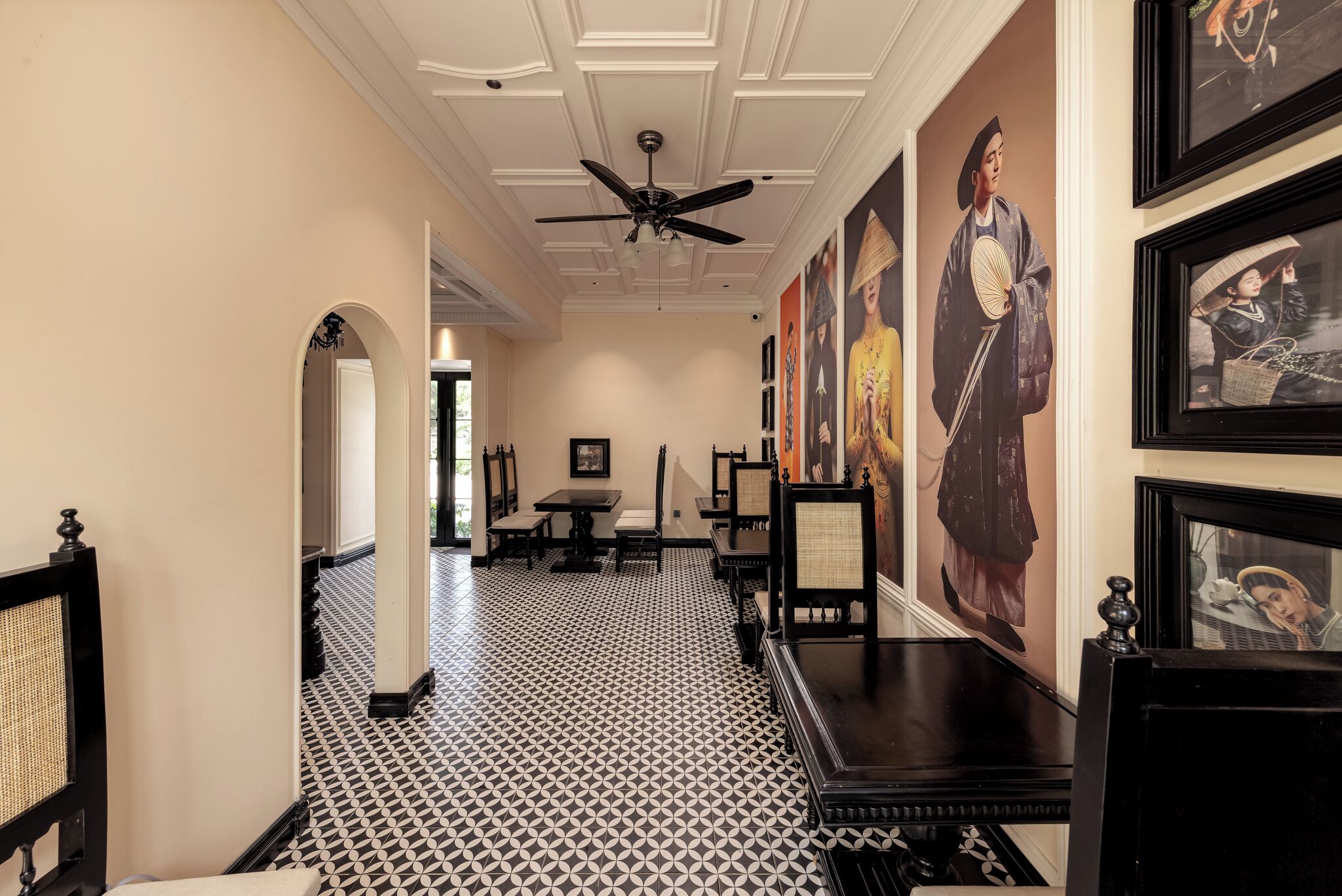 A hallway with black and white patterned flooring, a ceiling fan, framed artwork on the walls, and a dining area with tables and chairs.
