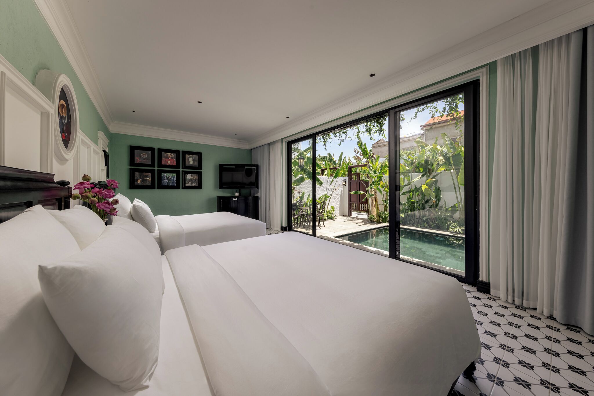 A hotel room with a large bed, a TV, a small table with flowers, and a view of a swimming pool and greenery outside.